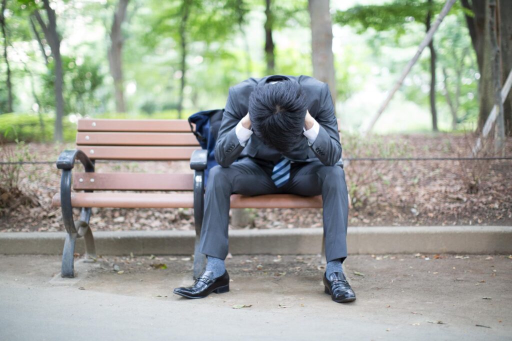 公園のベンチで頭を抱えうつむいているスーツ姿の男性