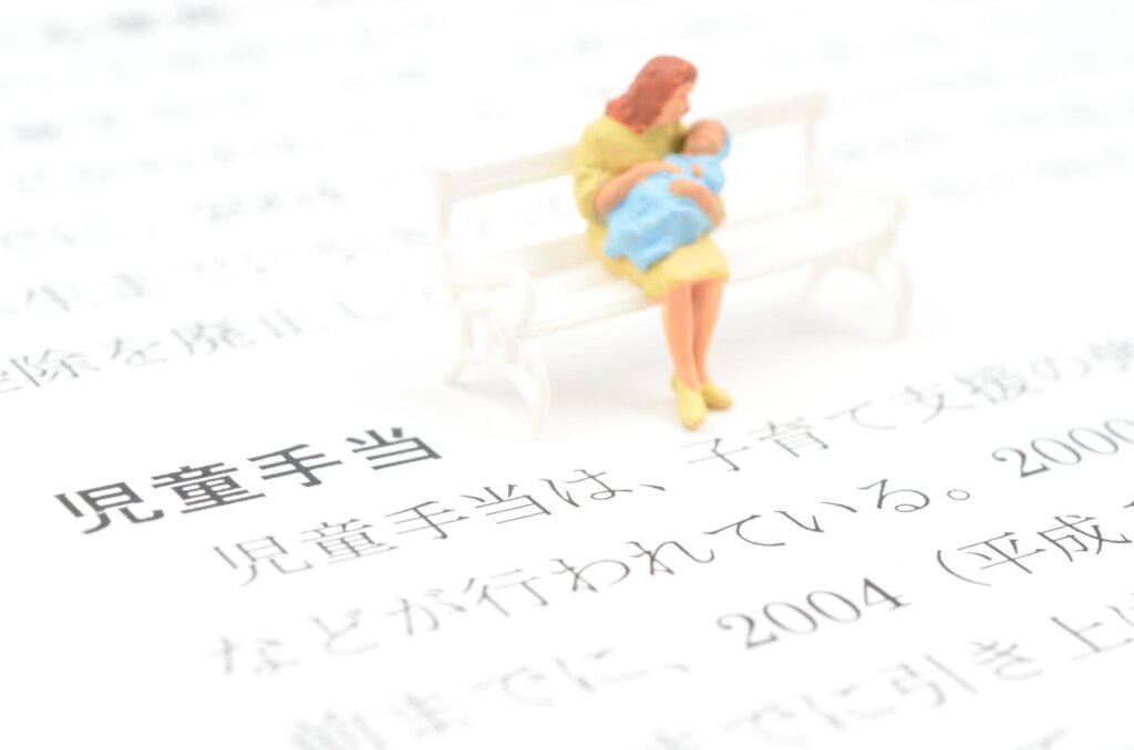 「児童手当」と書かれた書類の上に、ベンチに座って赤ちゃんを抱える母親のミニチュアフィギュアが置かれている