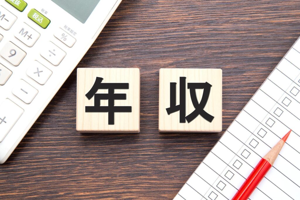 「年収」の漢字が書かれた木製ブロックと、電卓・チェックリスト・赤鉛筆が並ぶデスクの上のイメージ