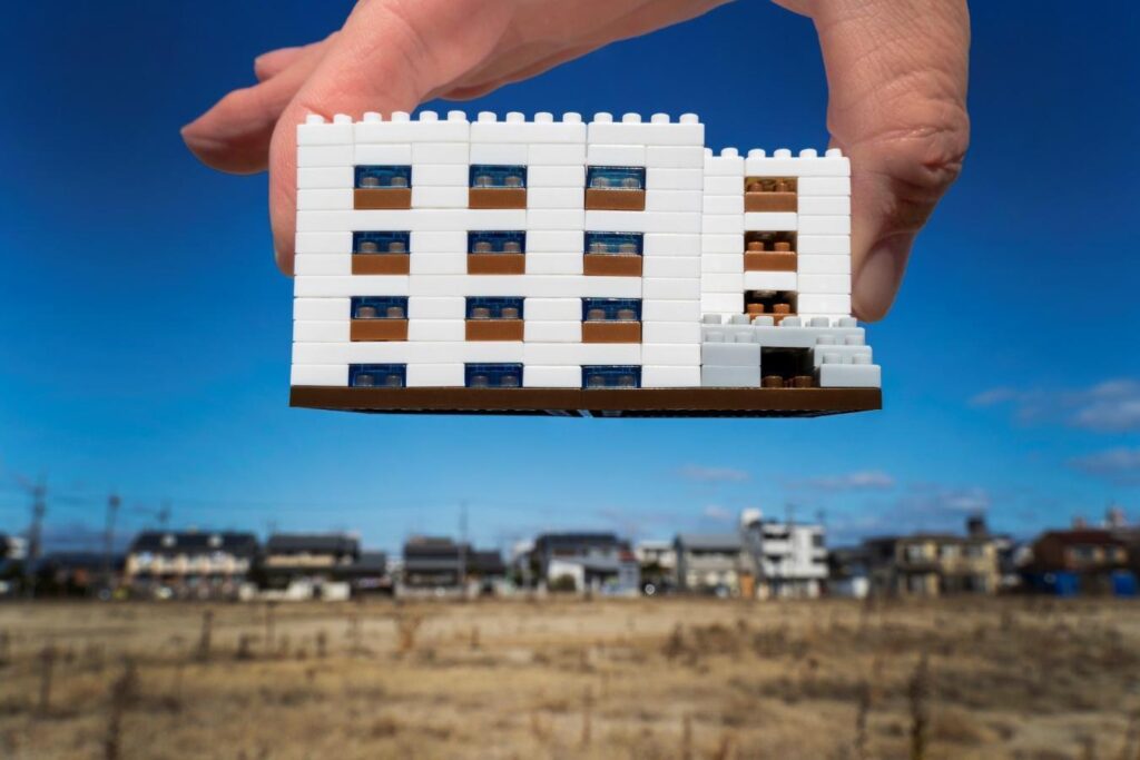 広がる空き地と空を背景に、ブロックでできた集合住宅を持つ人の手