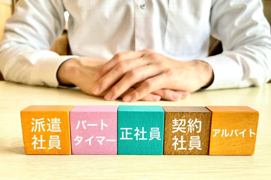 雇用形態を示す「派遣社員」「パートタイマー」「正社員」「契約社員」「アルバイト」の文字が書かれたカラフルな積み木と、手を組んで座る人