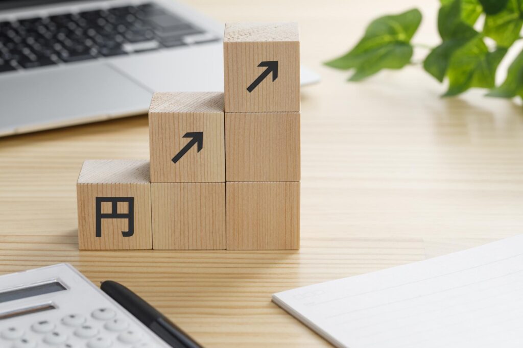 円マークと矢印が描かれた木製ブロックが階段状に積まれ、税負担や物価の上昇をイメージさせる構図