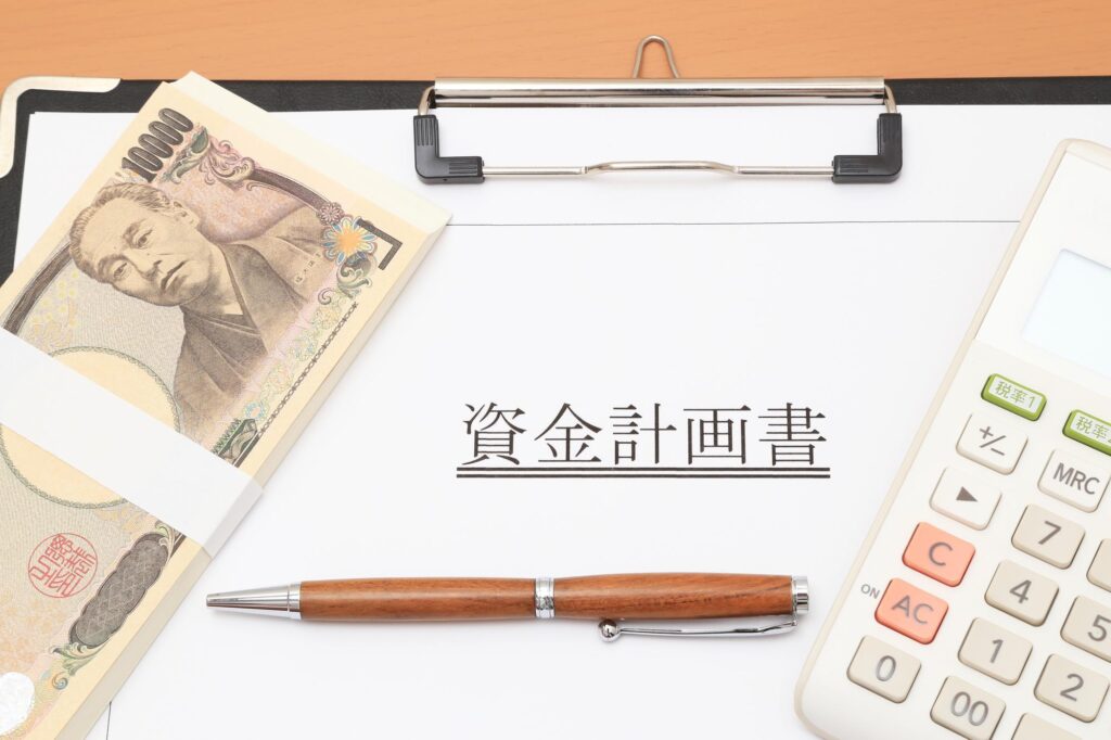 資金計画書と一万円札の束、電卓、ボールペンが並んだ机の上の書類